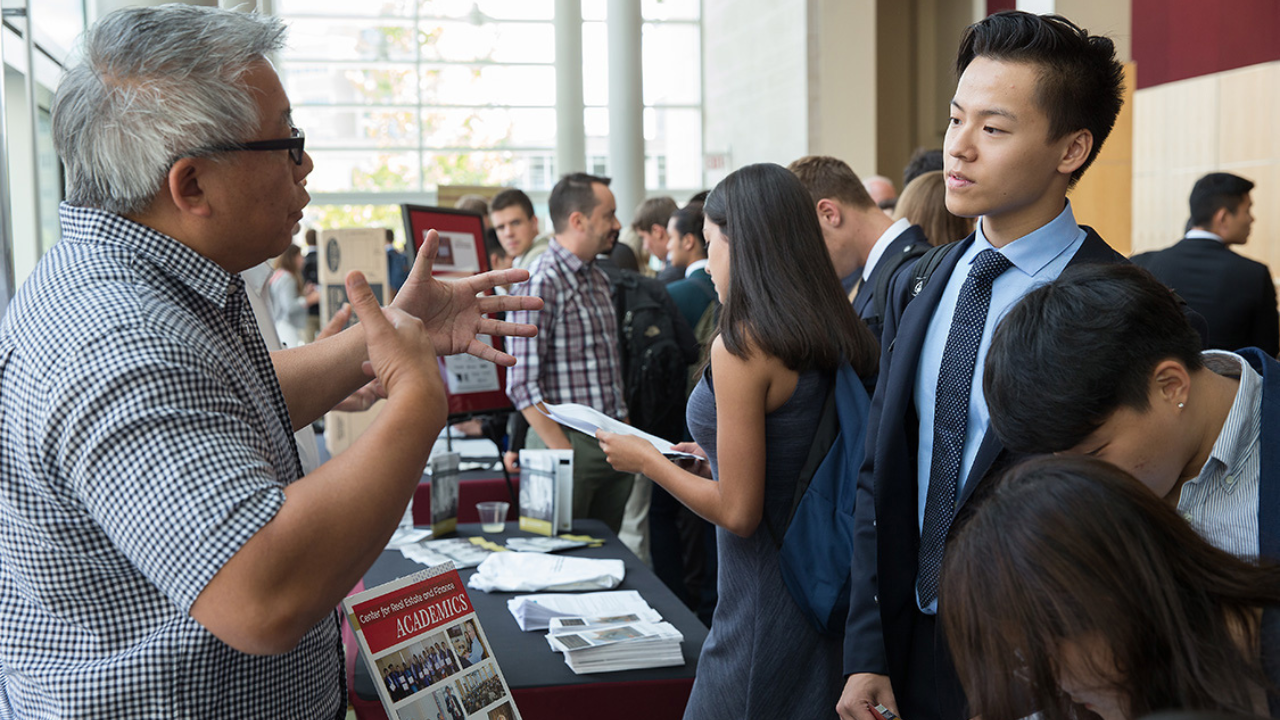 A man at a fair talks to a student dressed in a suit. 